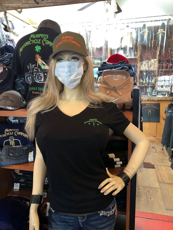 Female mannequin in a motorcycle shop wearing a mask and a black T-shirt with a green Murphys Motorcycle Company logo, standing beside a display of casual motorcycle clothing and accessories.