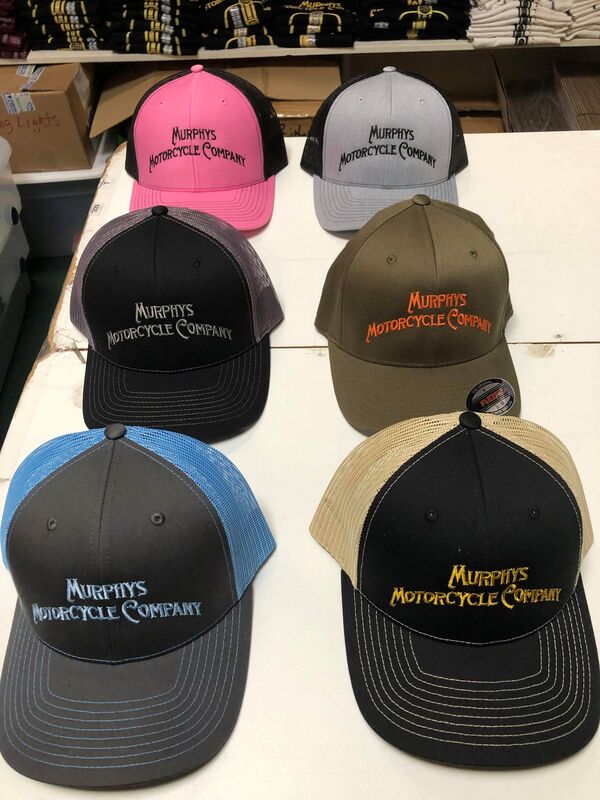 Collection of baseball caps in earth tones with the Murphy's Motorcycle Company logo showcased on a store wall.